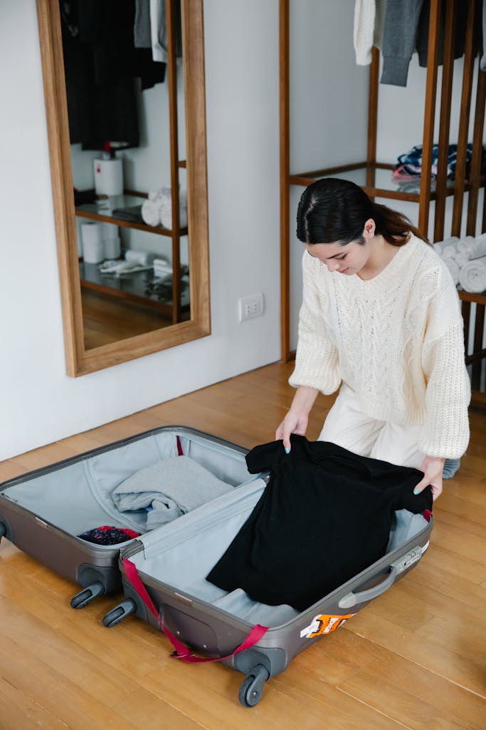 Woman kneeling on wooden floor packing clothes into luggage for travel preparation.